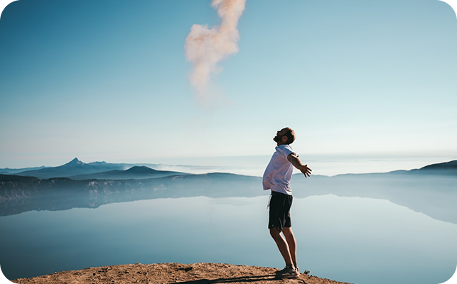 Person standing on a cliff with arms outstretched, looking towards the horizon and smoke rising