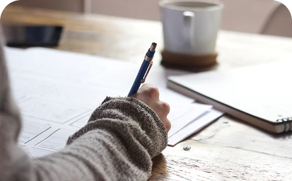 Person's hand holding a pen over papers on a wooden table with a cup in the background