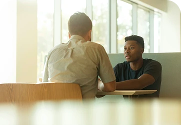 Two men sit at a table facing each other, engaged in conversation by a window