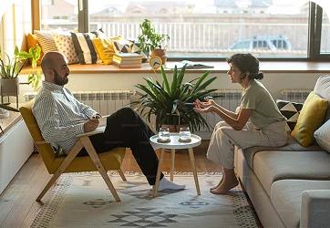 Two people sit in a bright living room, having a conversation over a small table