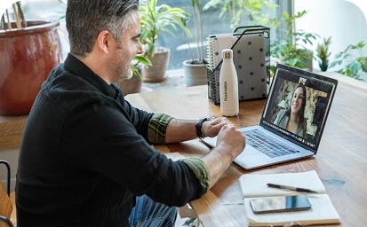A man video calls a woman on a laptop at a wooden table with plants around
