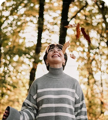 A person in a striped sweater smiles while standing among trees, surrounded by falling leaves