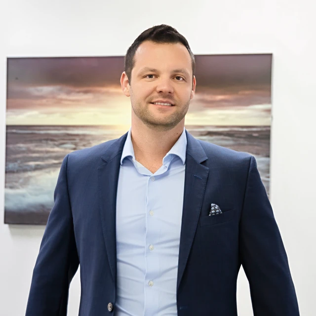 Man in a suit smiling in front of a landscape painting with ocean and sky